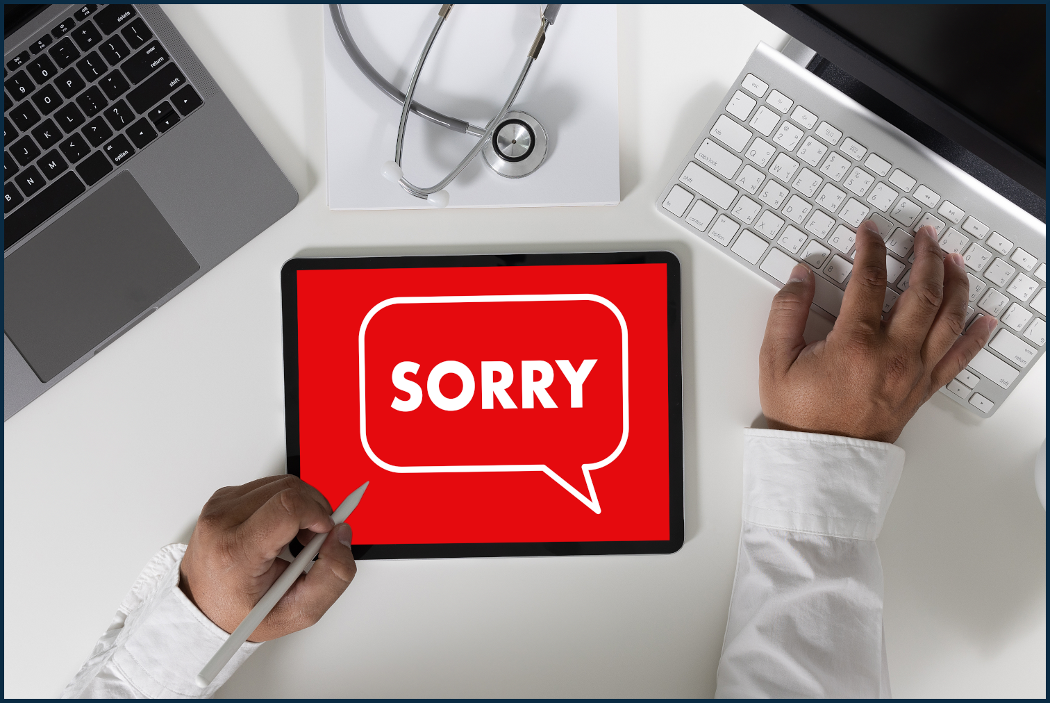 A desk with two hands resting on it, two laptops, and a tablet that has a red screen with the word sorry in white