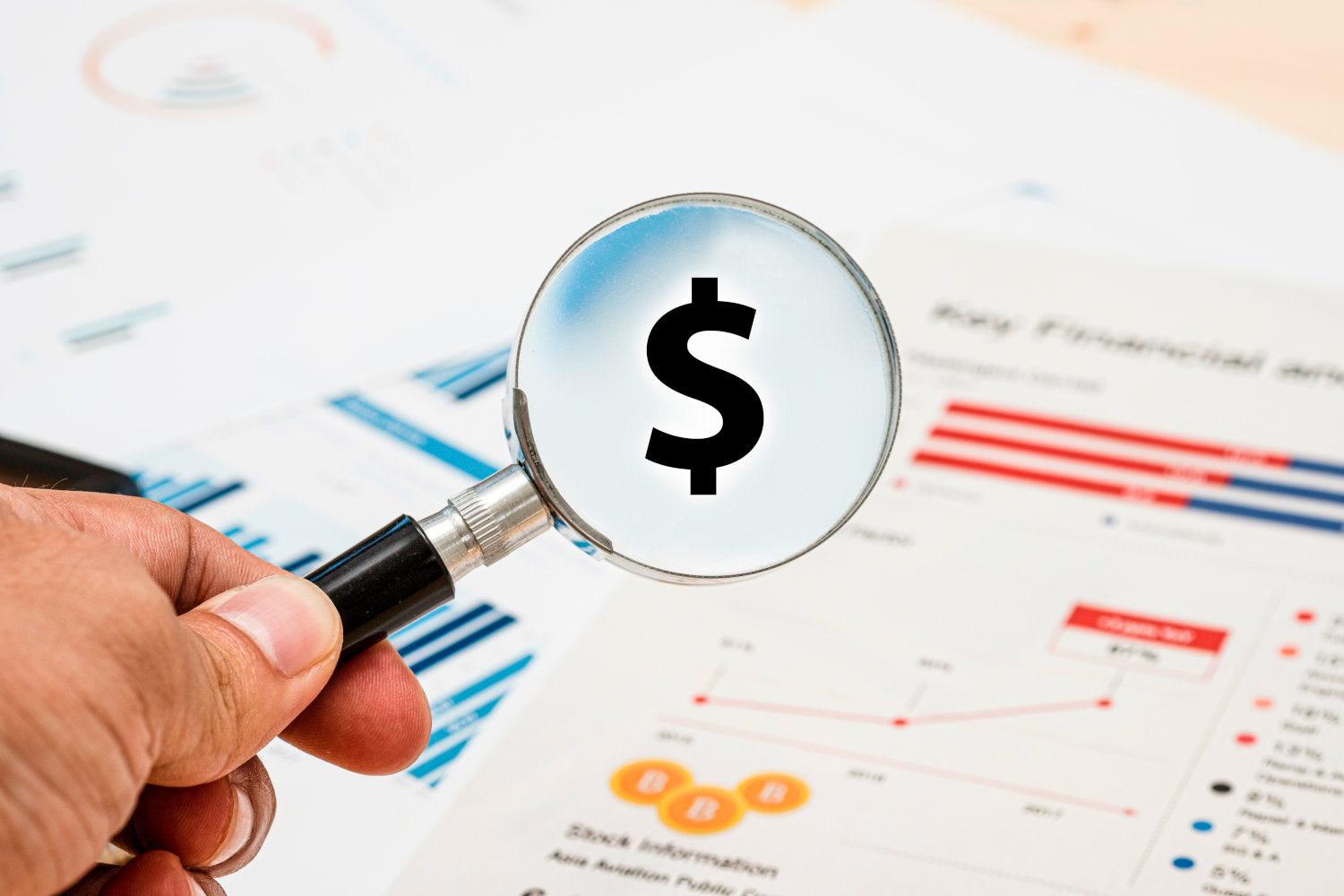 A mgnifying glass viewing a dollar sign with charts on a table in the background